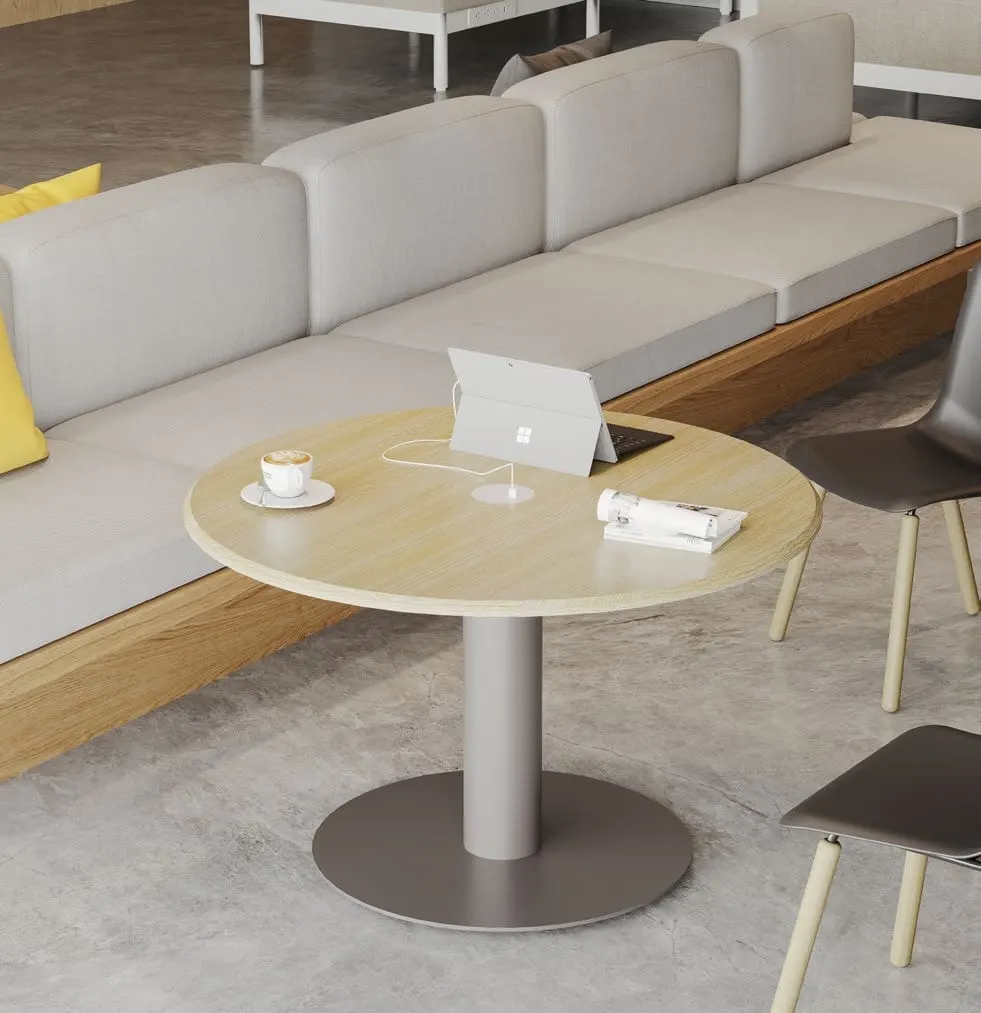 Round light-oak occasional table on a metal column base with laptop and power module beside a grey sofa